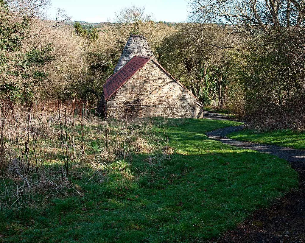The stone gable and conical chimney of Derwentcote Steel Furnace beyond rough grass and a curving path