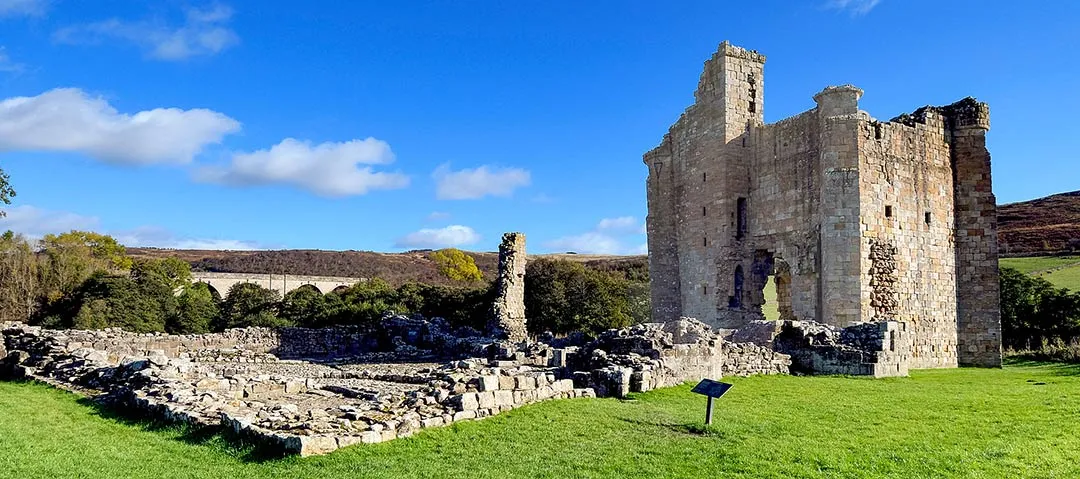 The ruined solar tower of Edlingham Castle above the courtyard footings on a sunny autumn day, with a railway viaduct beyond
