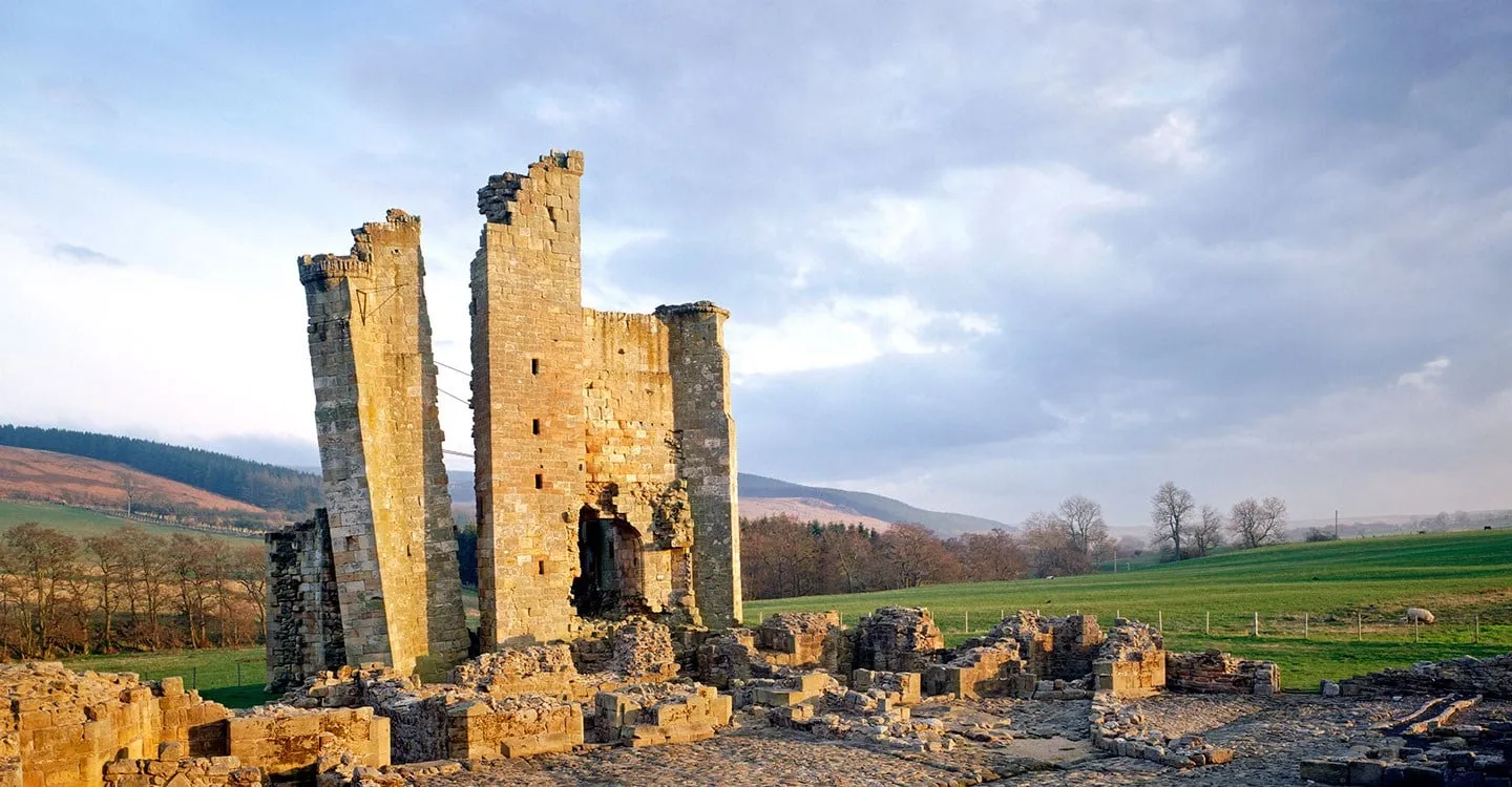 The leaning gatehouse wall and solar tower of Edlingham Castle glowing in low winter sun, with moorland hills and a sheep in the field