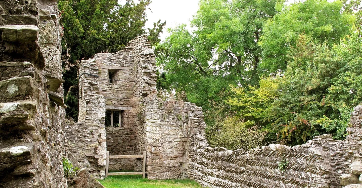 The ruined stone tower of Edvin Loach Old Church with a wooden stile, and herringbone masonry walls among green trees