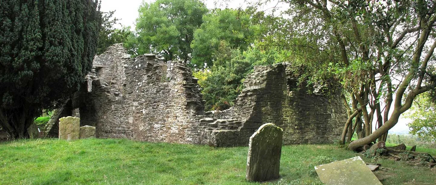 The ruined walls of Edvin Loach Old Church across a grassy churchyard with old headstones, a yew and leaning trees