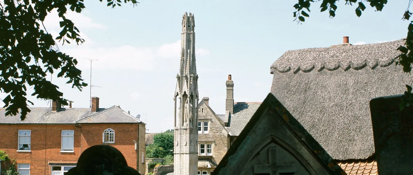 The slender spire of the Eleanor Cross rising above Geddington's brick and thatched rooftops, seen from the churchyard