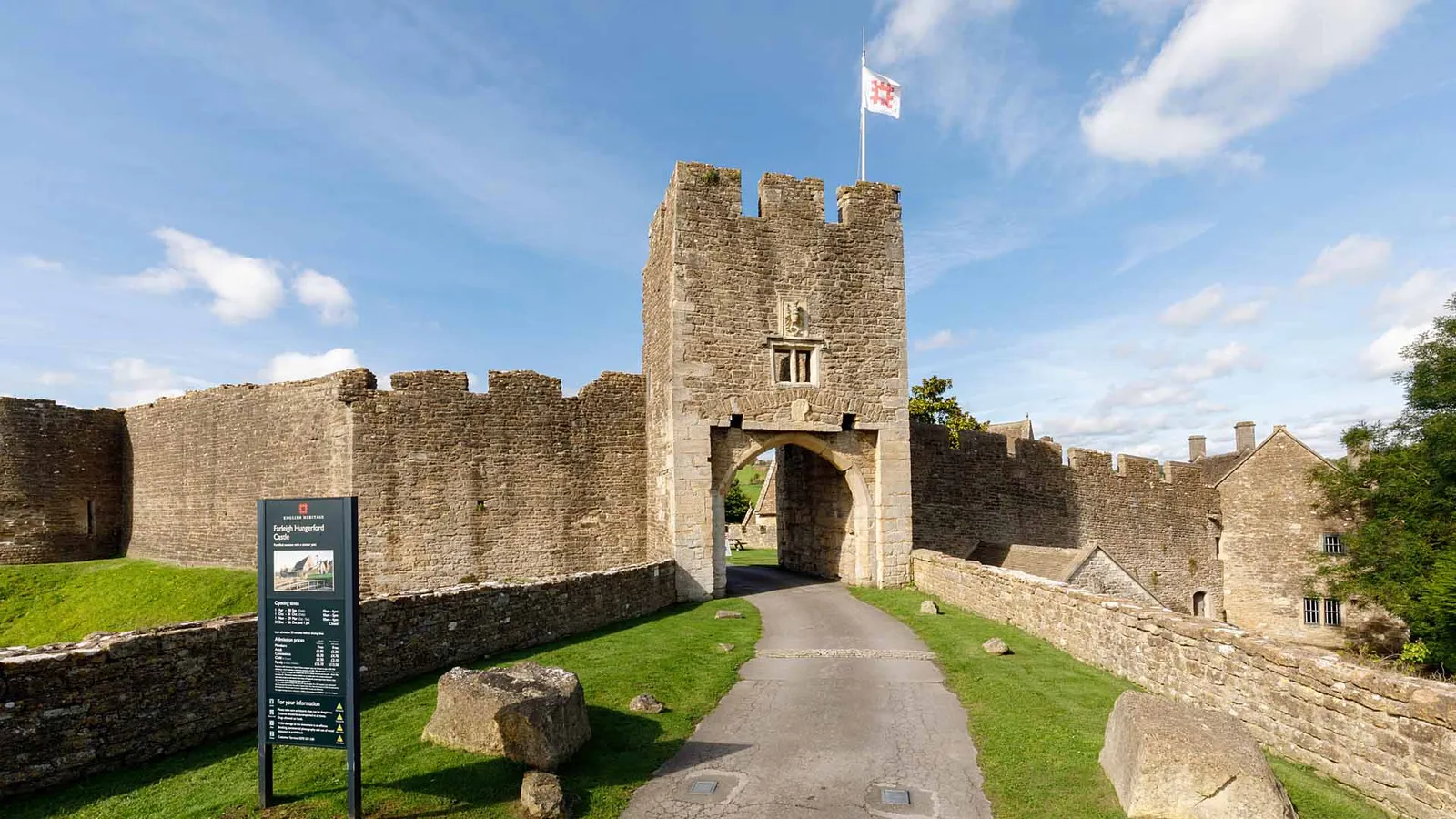The gatehouse of Farleigh Hungerford Castle with its flag, the entrance drive and curtain walls under a blue sky