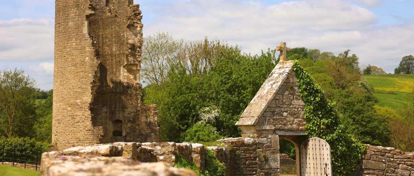 The ruined south-west tower of Farleigh Hungerford beside the ivy-covered churchyard gate, with fields beyond