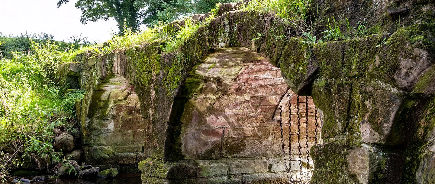 A moss-covered medieval stone bridge over a stream beside Furness Abbey, with an iron chain across its arch