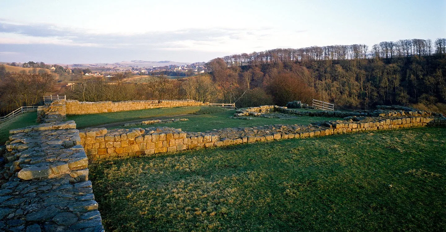 The low stone walls of Harrows Scar milecastle in golden evening light, with wooded slopes and a village in the valley beyond