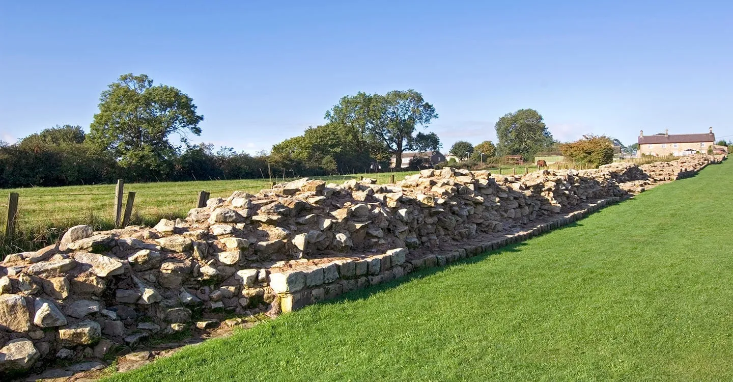 A long, broad stretch of Hadrian's Wall foundations beside a mown lawn, with fields, trees and a stone farmhouse beyond under a blue sky