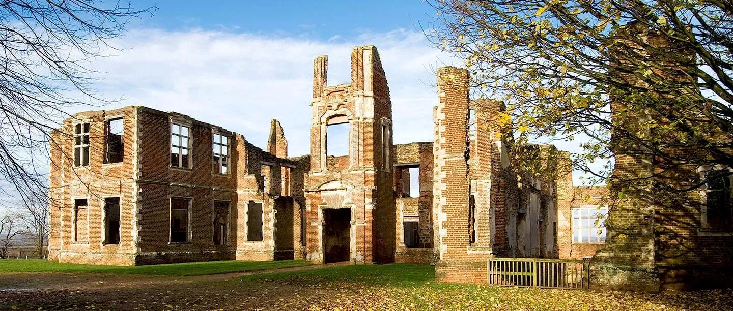 The roofless red-brick shell of Houghton House in autumn sunshine, with fallen leaves and a tree in golden leaf