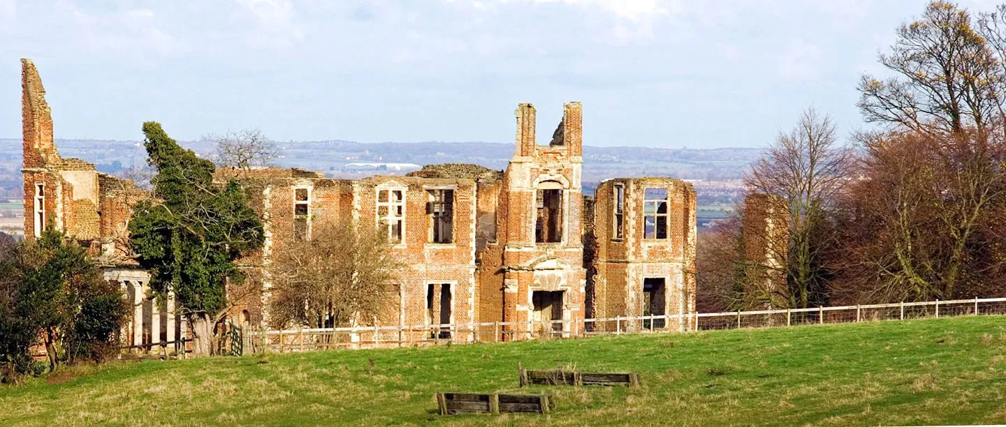 The ruined brick walls and towers of Houghton House above a grassy pasture, with the wide plain beyond