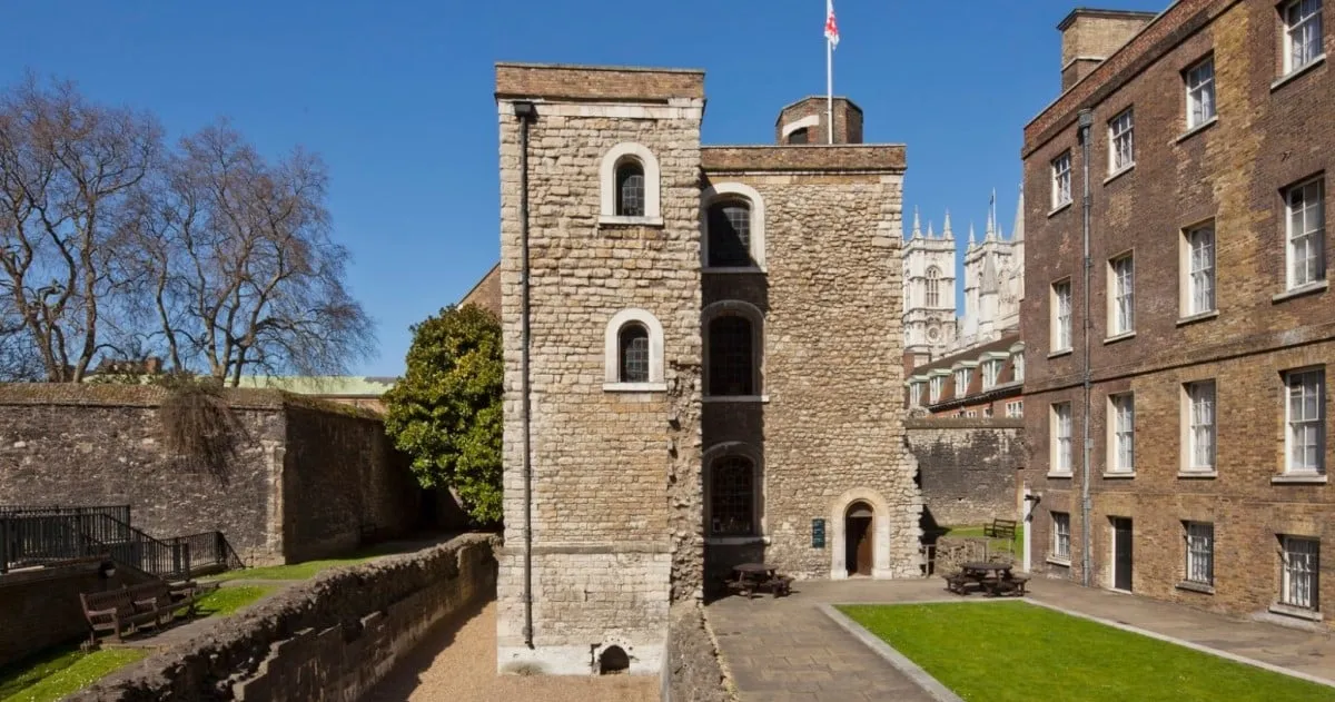 The stone Jewel Tower beside its medieval moat wall, with the towers of Westminster Abbey rising beyond, under a deep blue sky