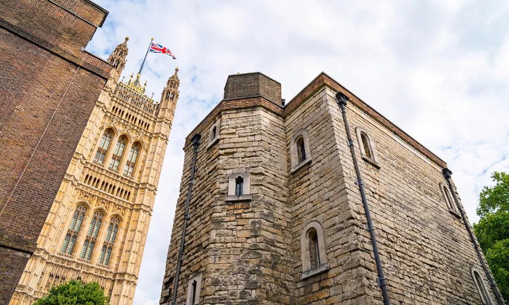 Looking up at the Jewel Tower's plain stone walls beside the ornate Victoria Tower of the Palace of Westminster, flying the Union flag