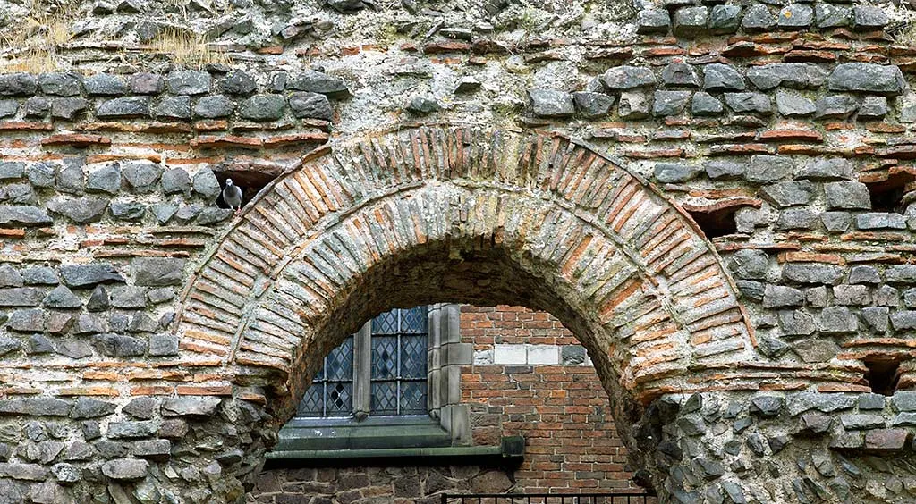 A Roman arch of thin red tiles set in the rubble of the Jewry Wall, framing a leaded church window beyond