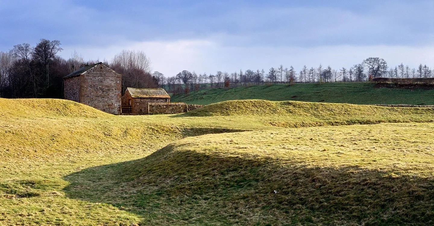 The grassy bank and ditch of King Arthur's Round Table henge in low winter sun, with stone field barns beyond