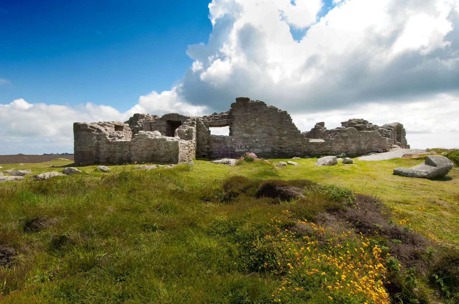 King Charles's Castle, roofless granite walls with an empty window, beyond heather and yellow gorse flowers under a towering cloud