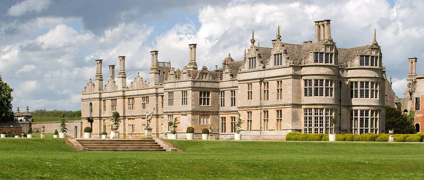 The south and west fronts of Kirby Hall with tall chimneys, a statue and garden steps under a cloudy sky