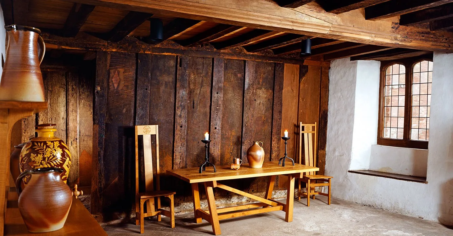 Inside Kirkham House, a medieval room with a plank-and-muntin screen, timber ceiling, oak table with candlesticks and a leaded window