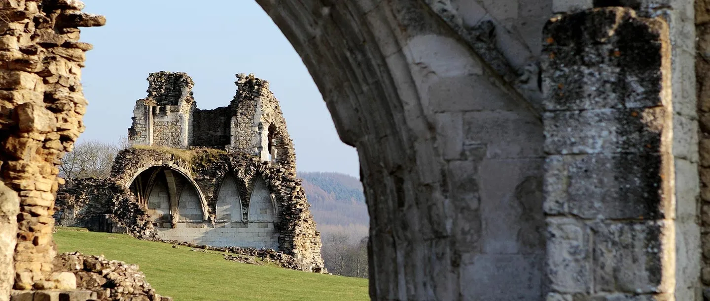 The vaulted lavatorium arches of Kirkham Priory seen through a ruined archway, with wooded hills beyond