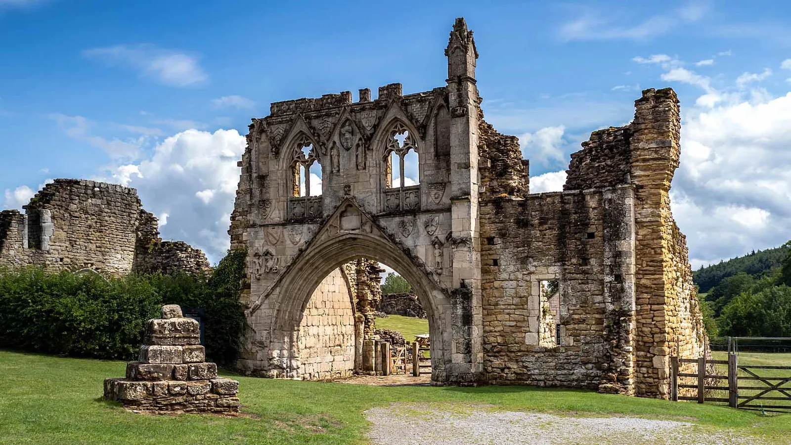 Kirkham Priory's carved gatehouse with its traceried windows and coats of arms, beside a stone cross base under a summer sky