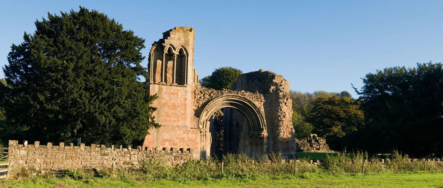 The ruined west front of Lilleshall Abbey with a round Norman arch and a tall arcaded wall, beside a large yew tree and a low wall