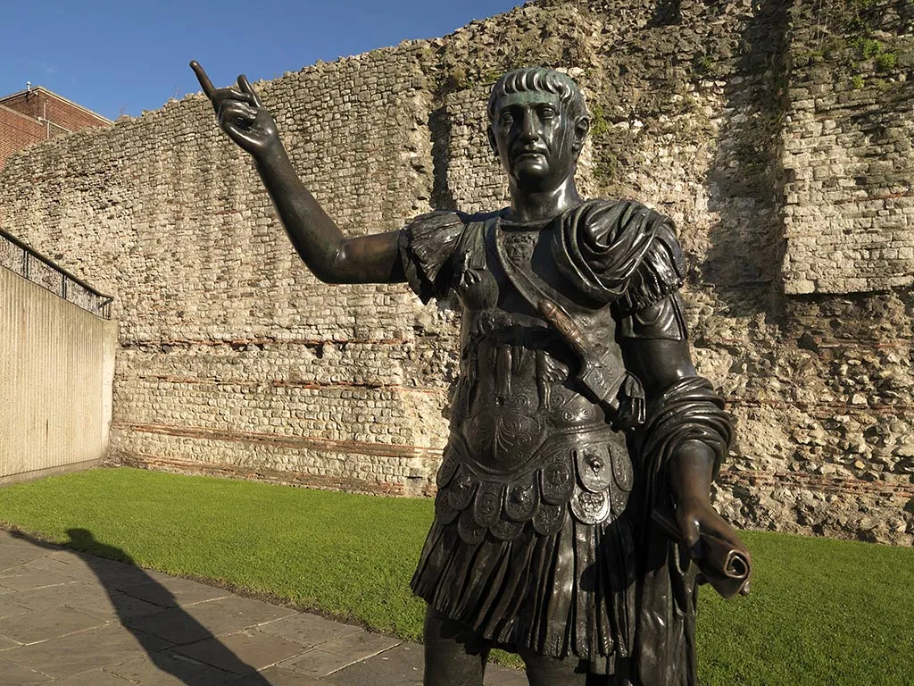 Bronze statue of the emperor Trajan in front of the Roman wall at Tower Hill, under a blue sky