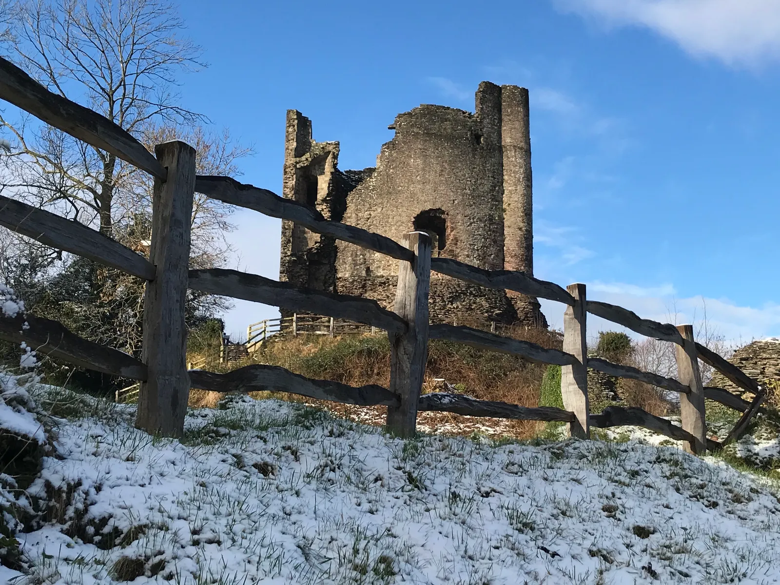 The ruined keep of Longtown Castle on its snow-covered motte behind a rustic wooden fence, under a bright blue sky