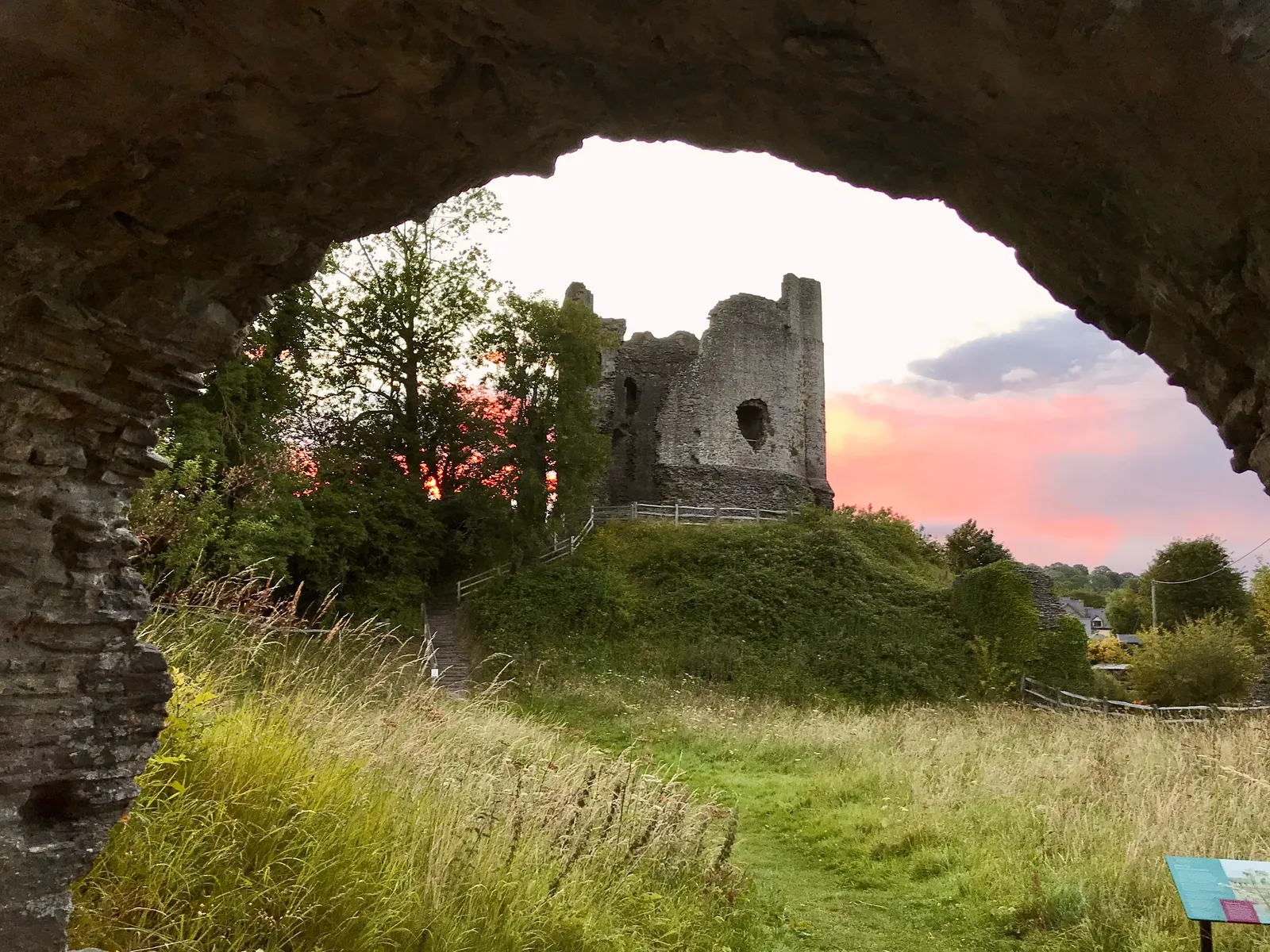 The keep of Longtown Castle on its motte at sunset, seen through a stone arch across a meadow of long grass under a pink sky