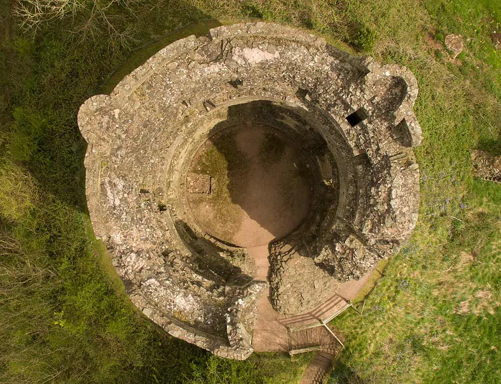 Looking straight down into the round keep of Longtown Castle, its thick walls and earth floor surrounded by grass and bluebells