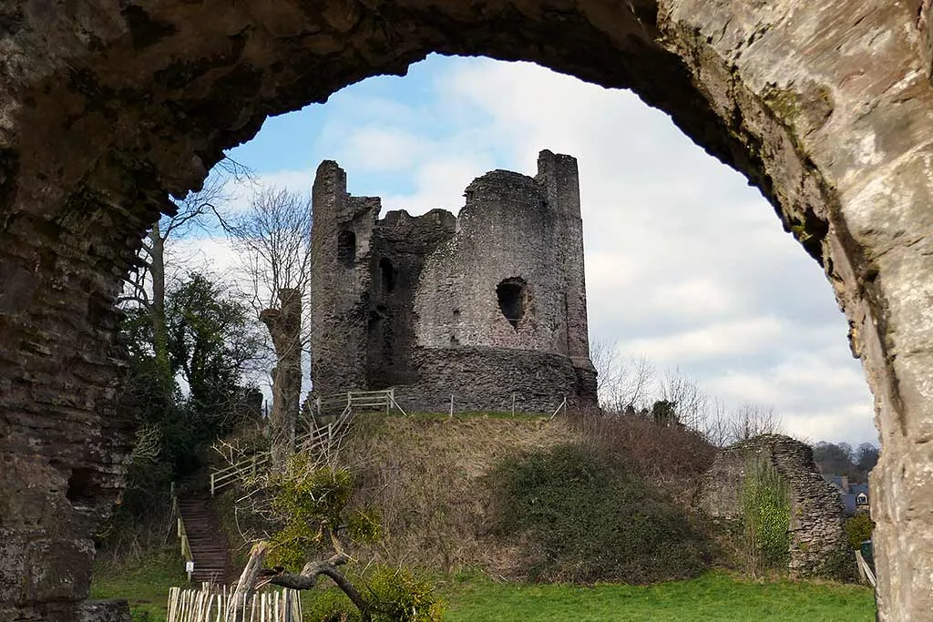 The round keep of Longtown Castle on its grassy motte with a wooden stair, framed by a rough stone archway