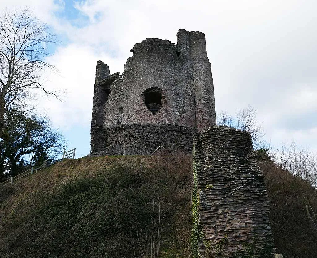 The cylindrical keep of Longtown Castle with a gaping window, high on its overgrown motte beside a stub of wall, under a cloudy sky