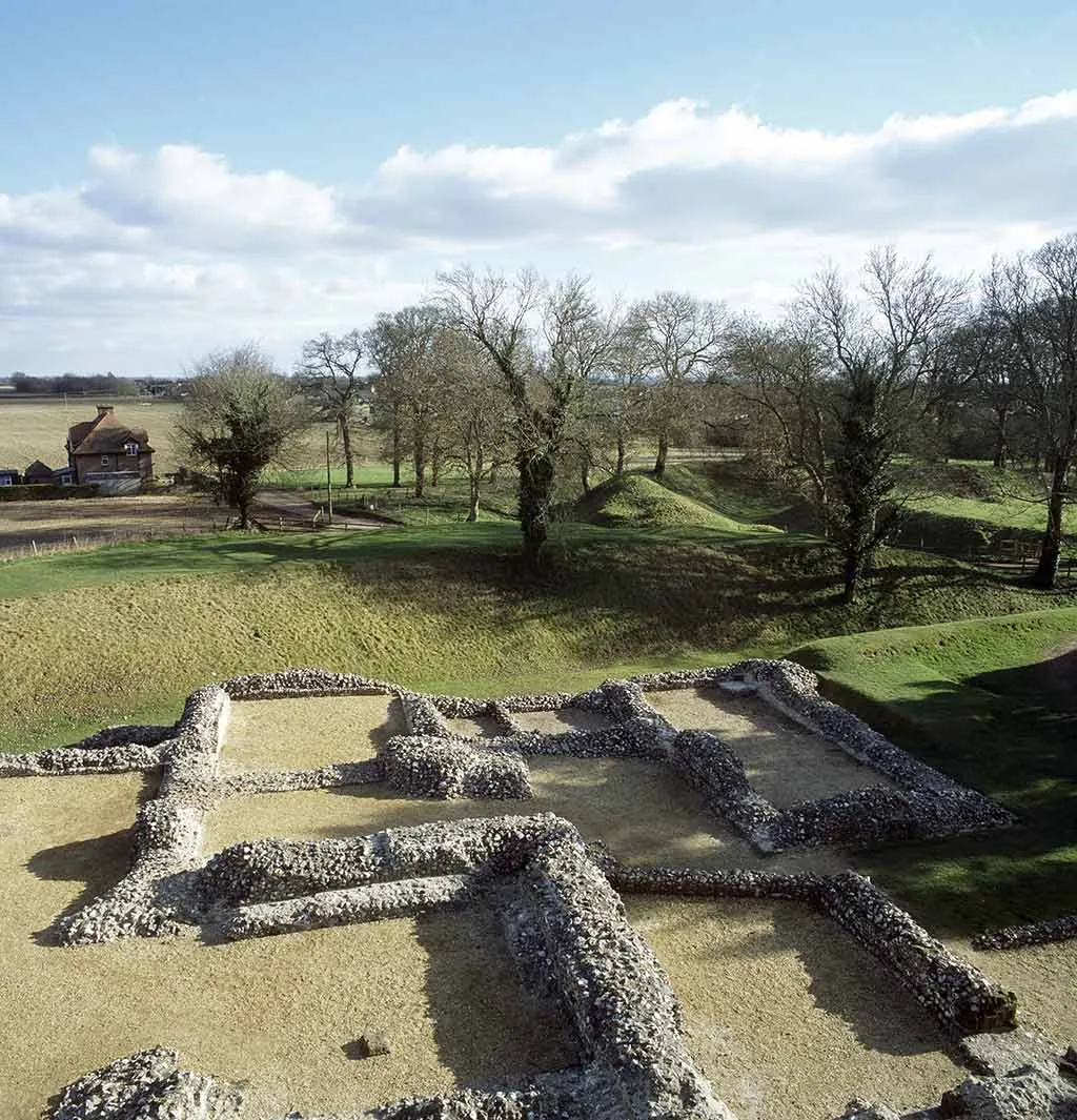 Flint wall foundations of the royal apartments at Ludgershall Castle on gravel, overlooking grassy earthworks, bare trees and a farmhouse