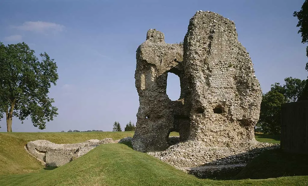 The tall flint ruin of the tower at Ludgershall Castle with a gaping window opening, beside low walls and grassy banks under a blue sky