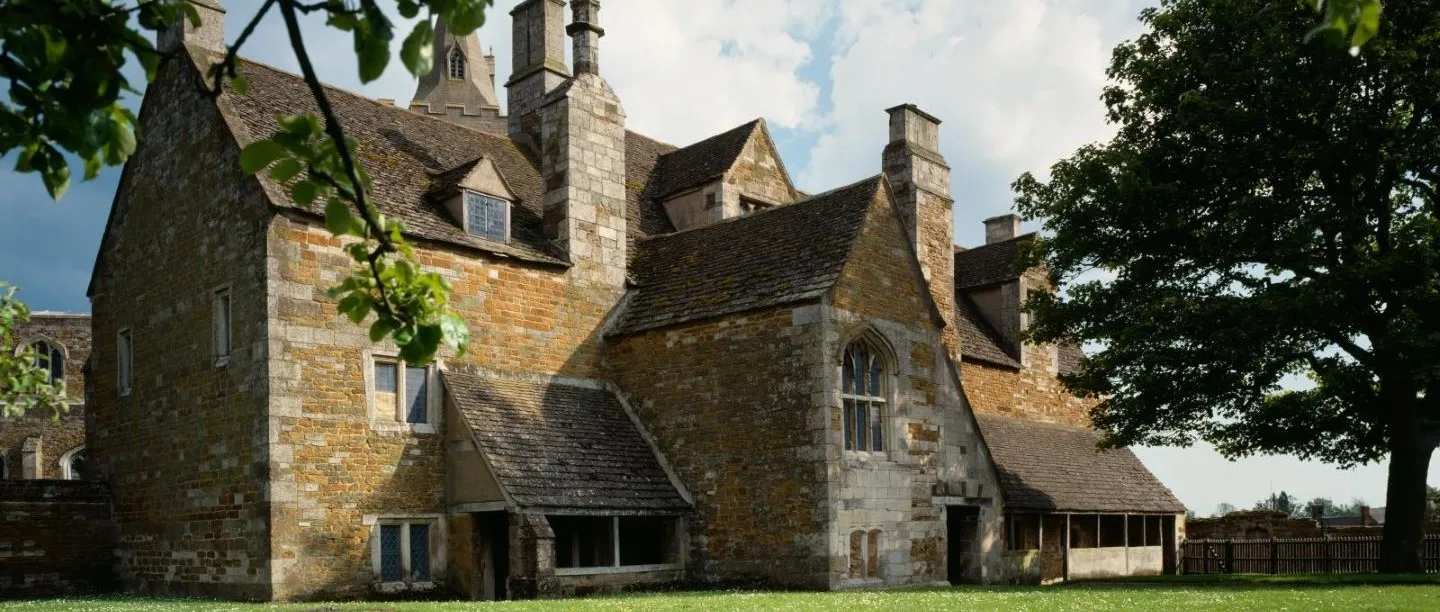 Lyddington Bede House, a long medieval ironstone building with tall chimneys, dormers and an open-fronted veranda, beside a lawn and an oak tree