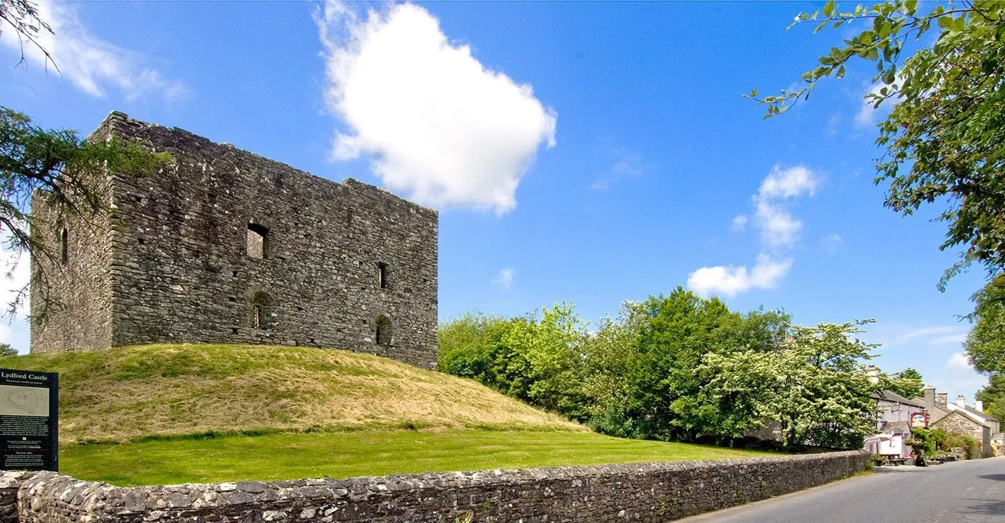 The ruined stone keep of Lydford Castle on its grassy mound beside the village road under a blue summer sky