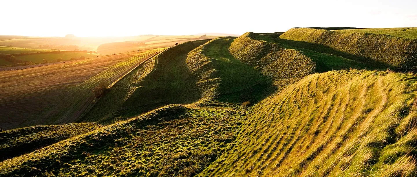Maiden Castle's grassy ramparts and ditches lit by low golden sun, with farmland beyond