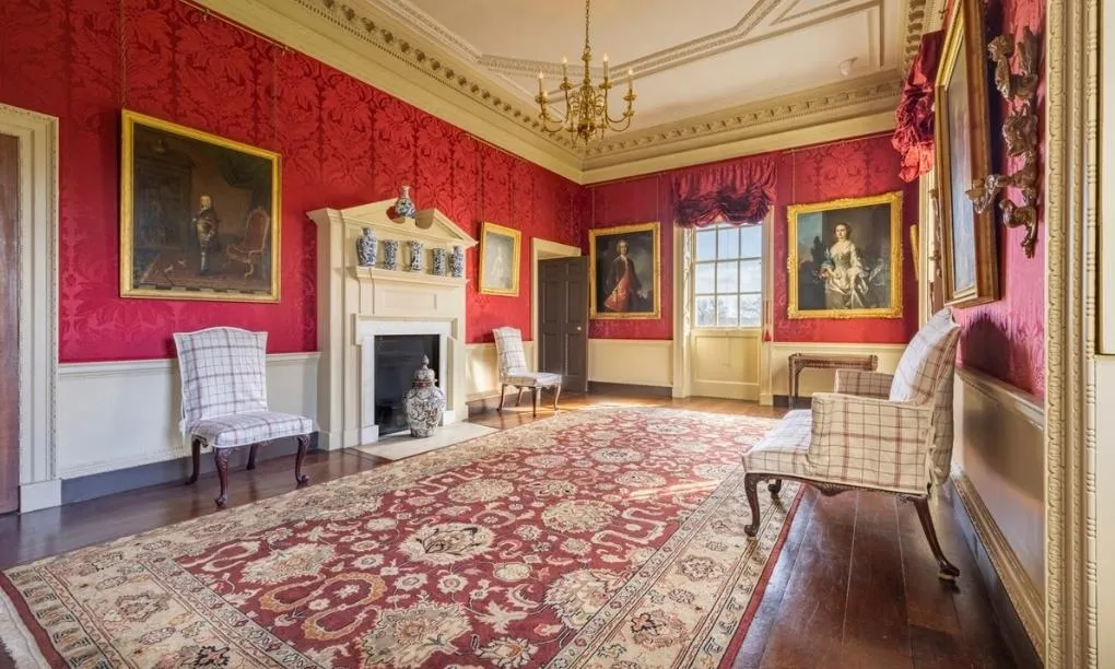 The Great Room at Marble Hill with red damask walls, gilt-framed portraits, a carved fireplace and checked chairs