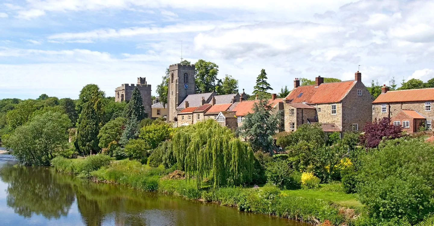Marmion Tower and the church tower above West Tanfield's stone cottages and gardens on the River Ure under a summer sky