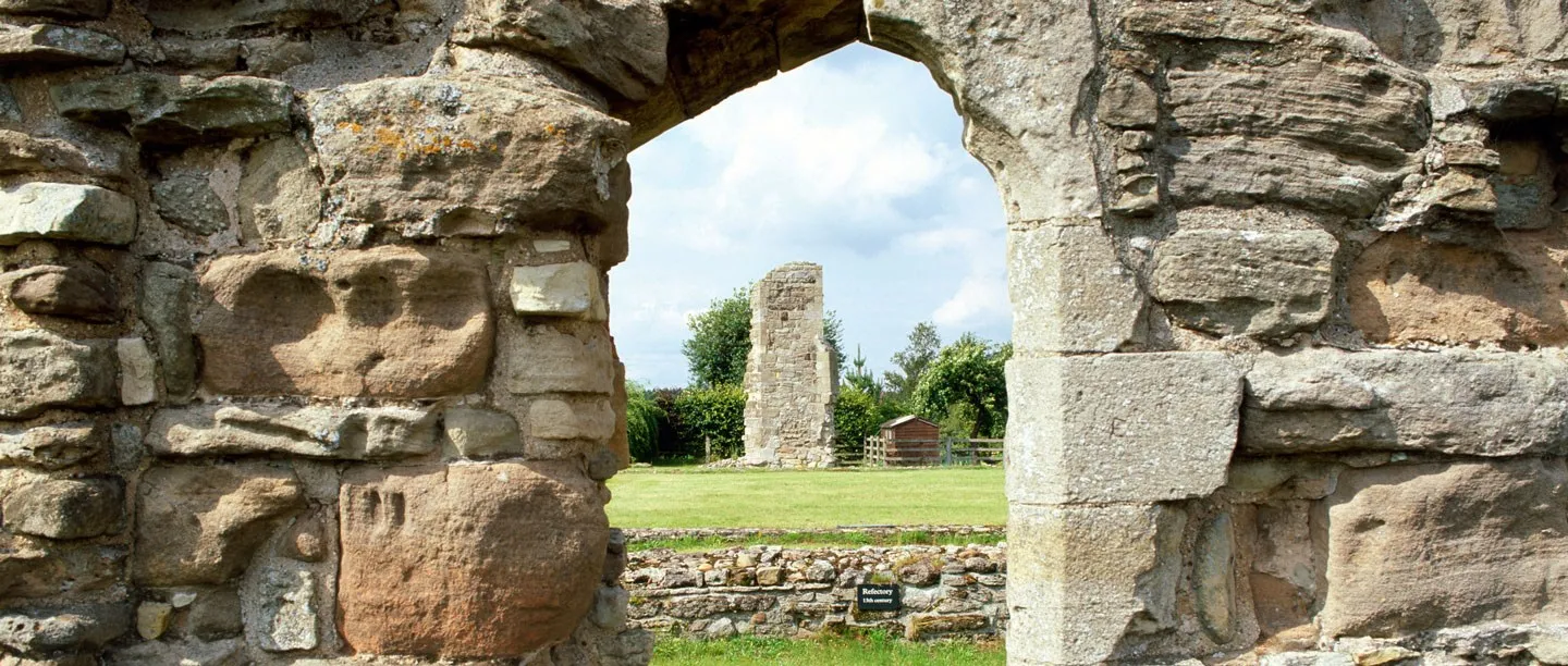 A view through a ruined stone doorway at Mattersey Priory to a tall fragment of the church tower on a mown lawn