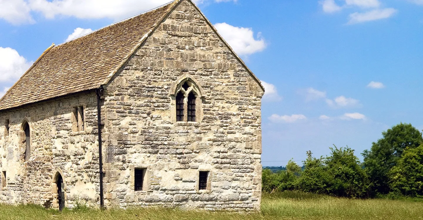 Meare Fish House, a medieval stone building with a steep tiled roof and a traceried gable window, in a summer meadow