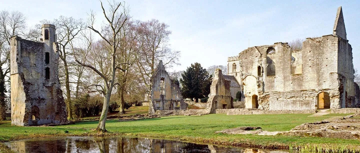 The ruined hall and tall tower of Minster Lovell Hall among bare trees, reflected in a pond