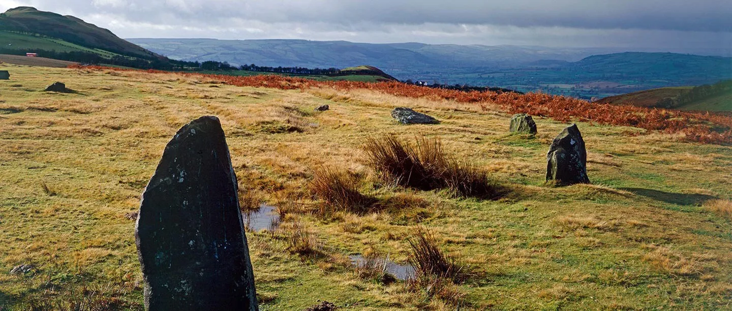 Weathered stones of Mitchell's Fold stone circle on moorland with rust-coloured bracken, looking over a wide valley under a stormy sky