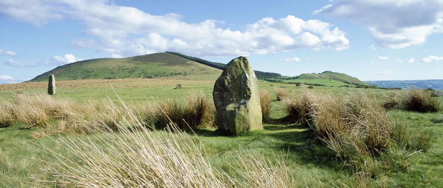 A tall lichen-covered standing stone of Mitchell's Fold among tussocky grass, with Corndon Hill and white clouds beyond