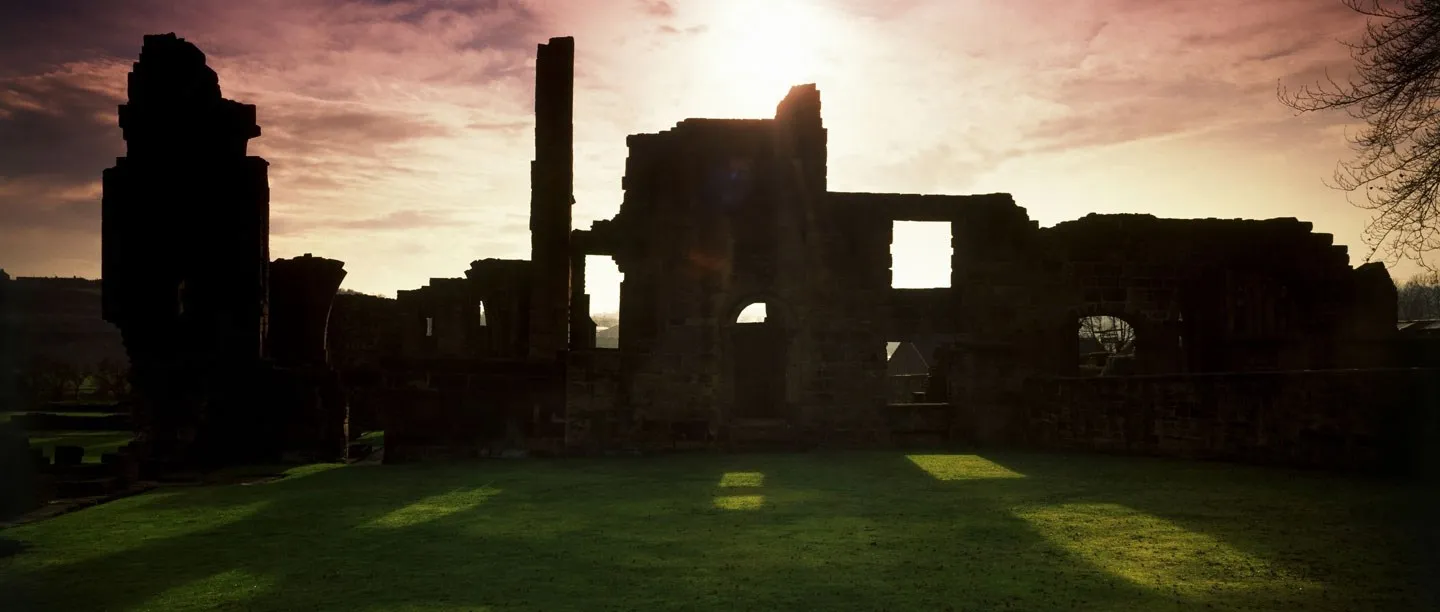 The ruins of Monk Bretton Priory silhouetted against a glowing dusk sky, sunlight streaming through window openings onto the lawn