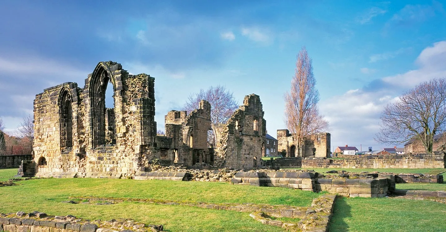 The ruined church and cloister of Monk Bretton Priory with a tall empty Gothic window, low walls on green lawns and the gatehouse beyond