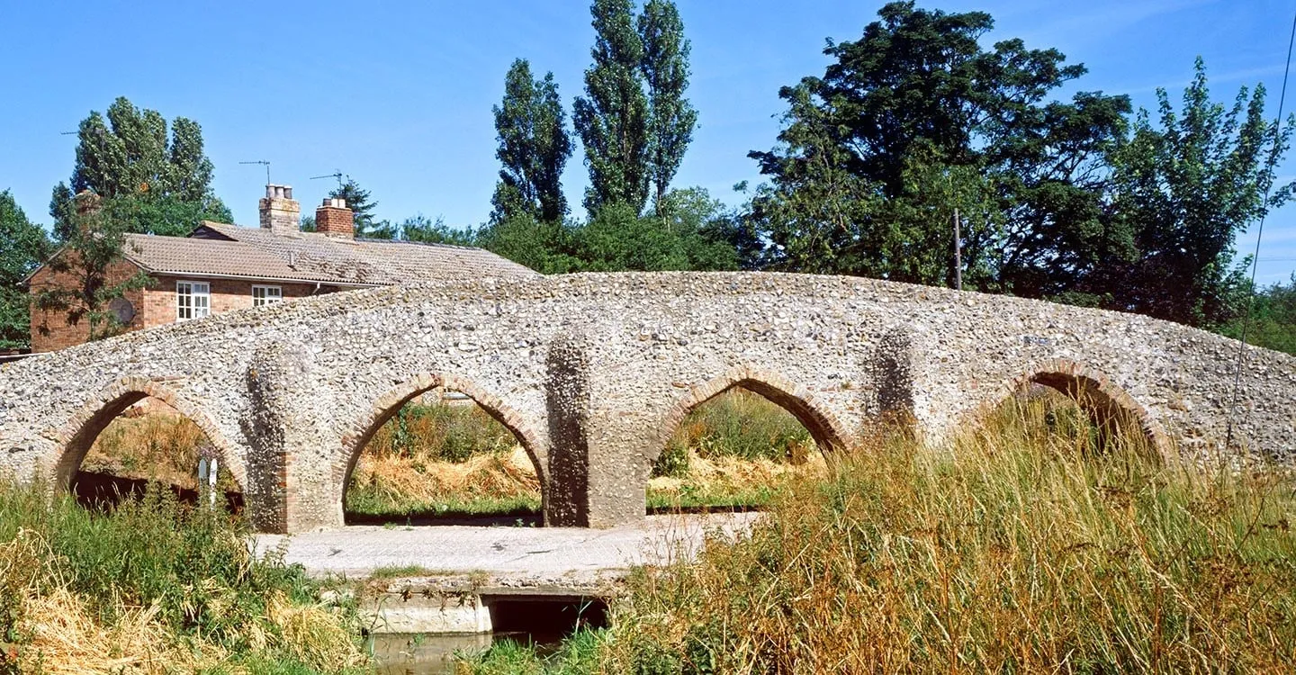 Moulton Packhorse Bridge, a medieval flint bridge with four pointed brick-edged arches over a shallow ford, on a summer day