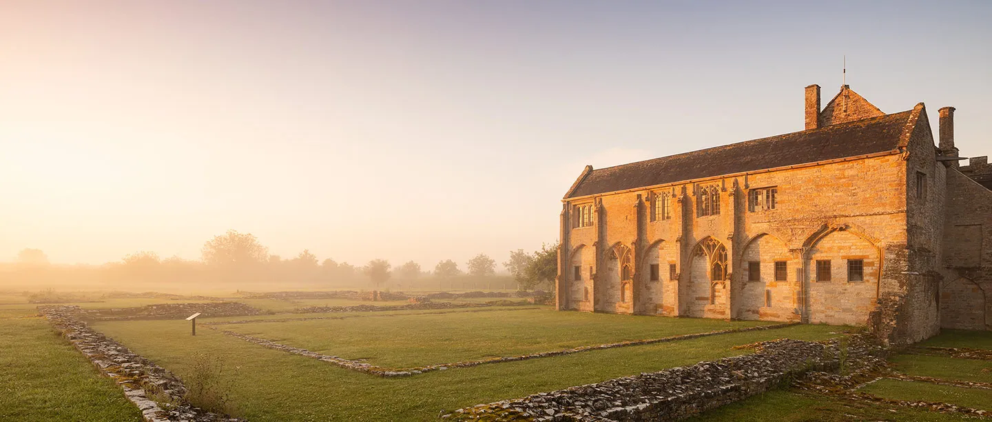 The cloister range of Muchelney Abbey glowing gold at sunrise, with mist over the excavated wall footings of the abbey church