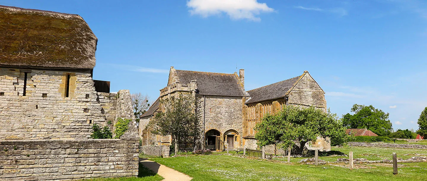 The thatched monks' privy and the abbot's house at Muchelney Abbey, with an apple tree, a bench and a path across a sunny lawn