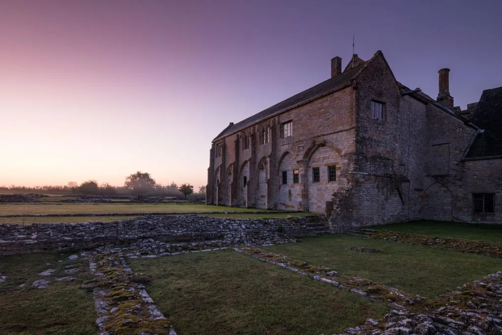 The cloister range of Muchelney Abbey at dusk under a violet sky, above the moss-covered footings of the abbey buildings