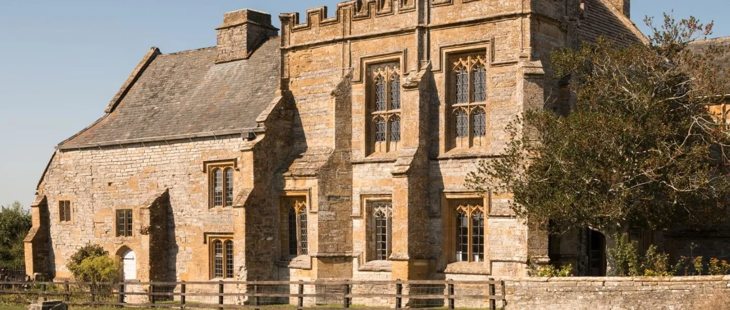 The battlemented abbot's house at Muchelney Abbey with tall traceried windows in golden stone, behind a wooden fence