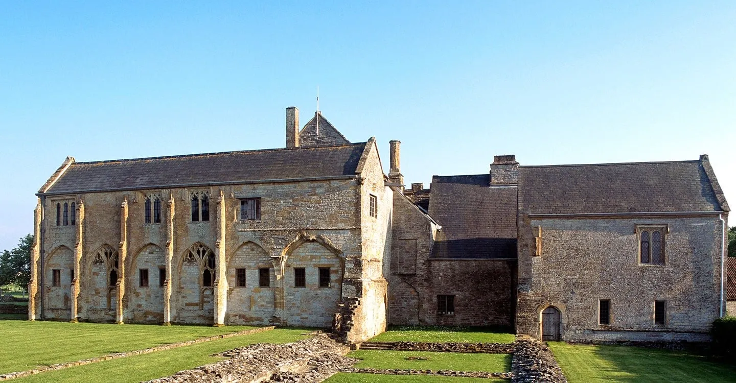 The long cloister range of Muchelney Abbey with its blind traceried arcade beside the abbot's house, above the excavated wall footings on a lawn in evening sun