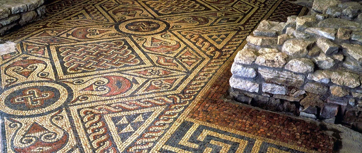 The intricate Roman mosaic floor at North Leigh Roman Villa, with interlaced knots and geometric patterns in red, cream and grey beside a rough stone wall